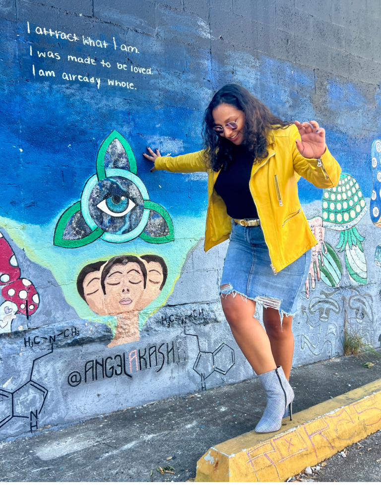 Woman wearing a Yellow Jacket and a jean skirt and boots against a mural about manifestation