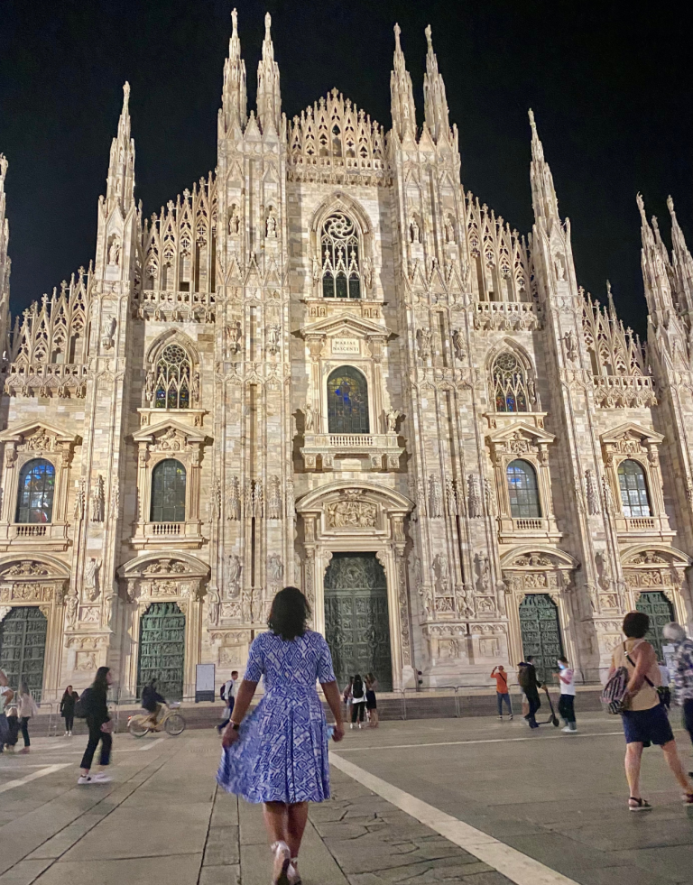 Mujer en un vestido blanco y azul parada frente al Duomo di Milano durante la noche.