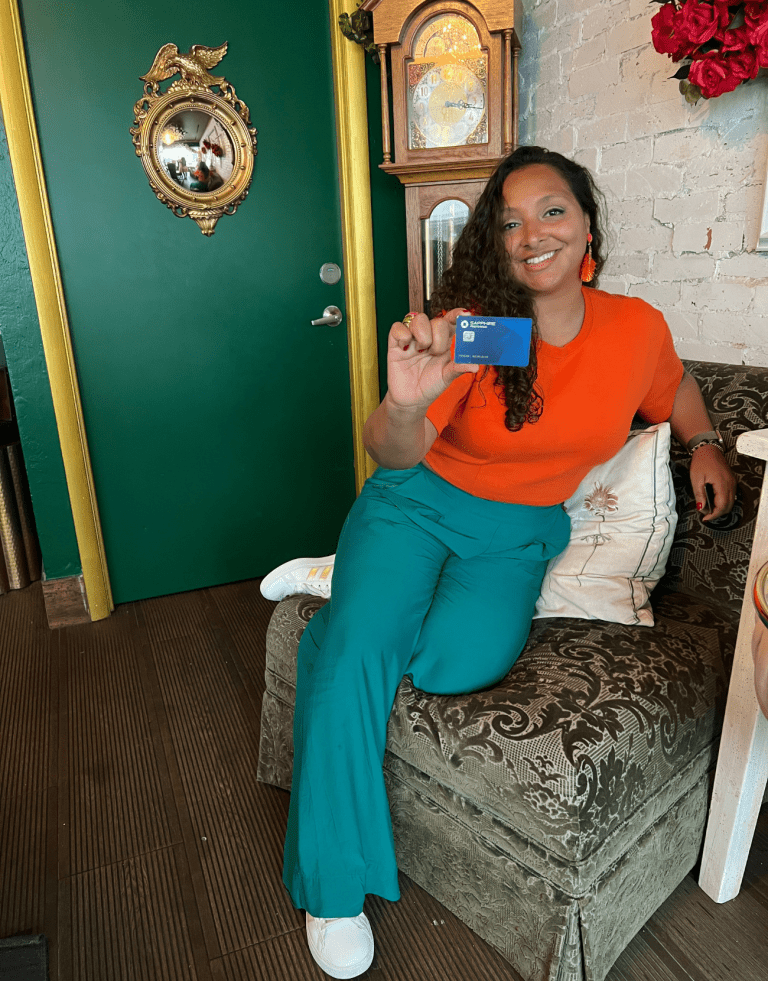 Latina holding up her credit card. She is wearing Teal and Orange pants while sitting in a coffee shop. This Latina woman is the author and she shares Credit Card Travel Hacking strategies to help you expand your travel budget