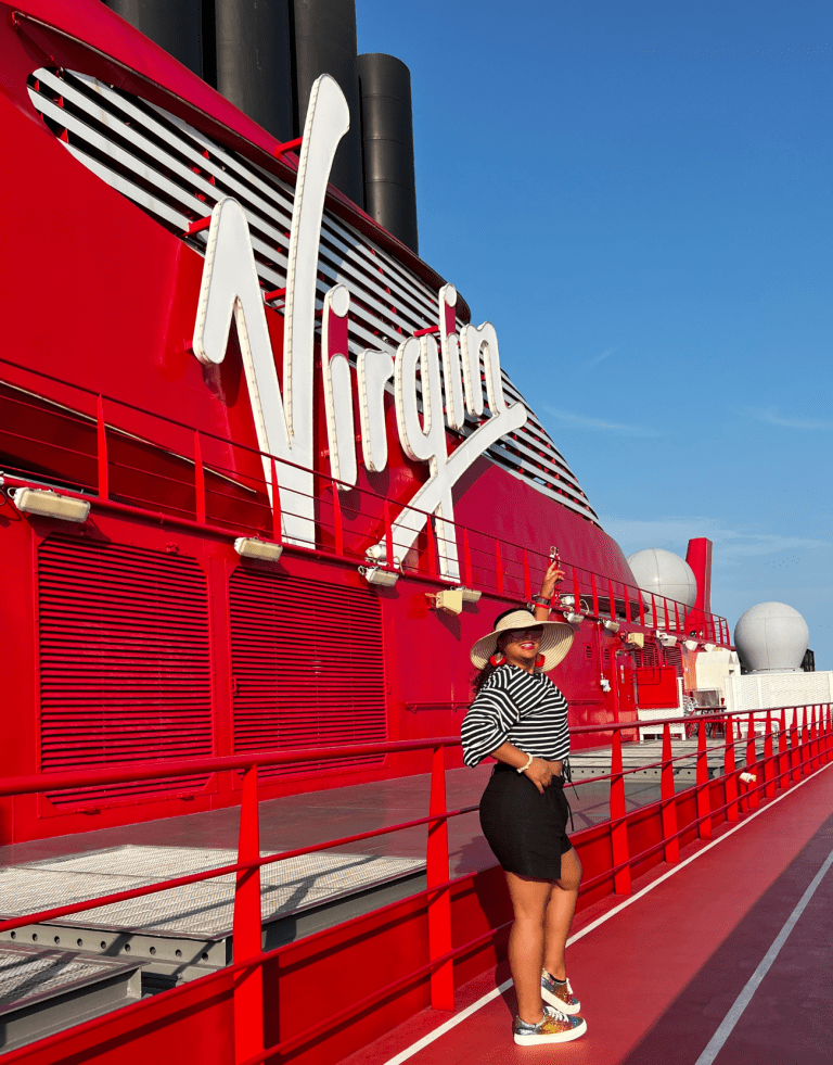 Una mujer latina vestida con ropa casual de resort, de pie en la cubierta de un barco de Virgin Voyages, mirando el océano con el diseño moderno del barco y los asientos al aire libre a la vista. La mujer es la autora y comparte Consejos Exclusivos para Virgin Voyages.