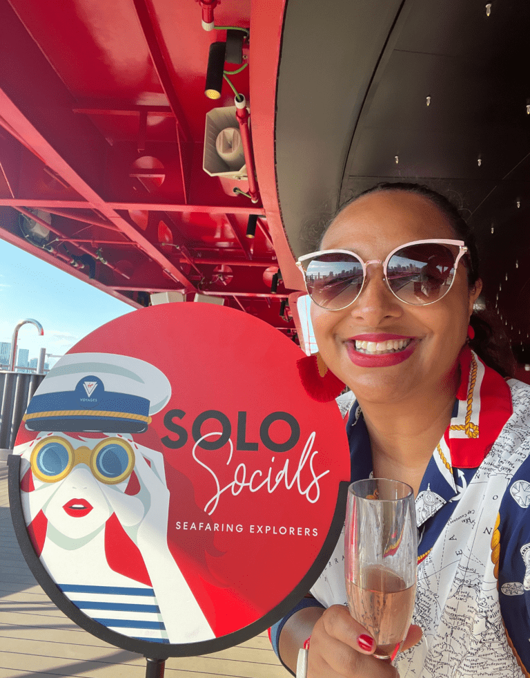 Viajera latina con un blusa nautica, posando junto al letrero de 'Solo Socials' de Virgin Voyages, sosteniendo una copa de champán y lista para conectarse con otros viajeros solitarios.