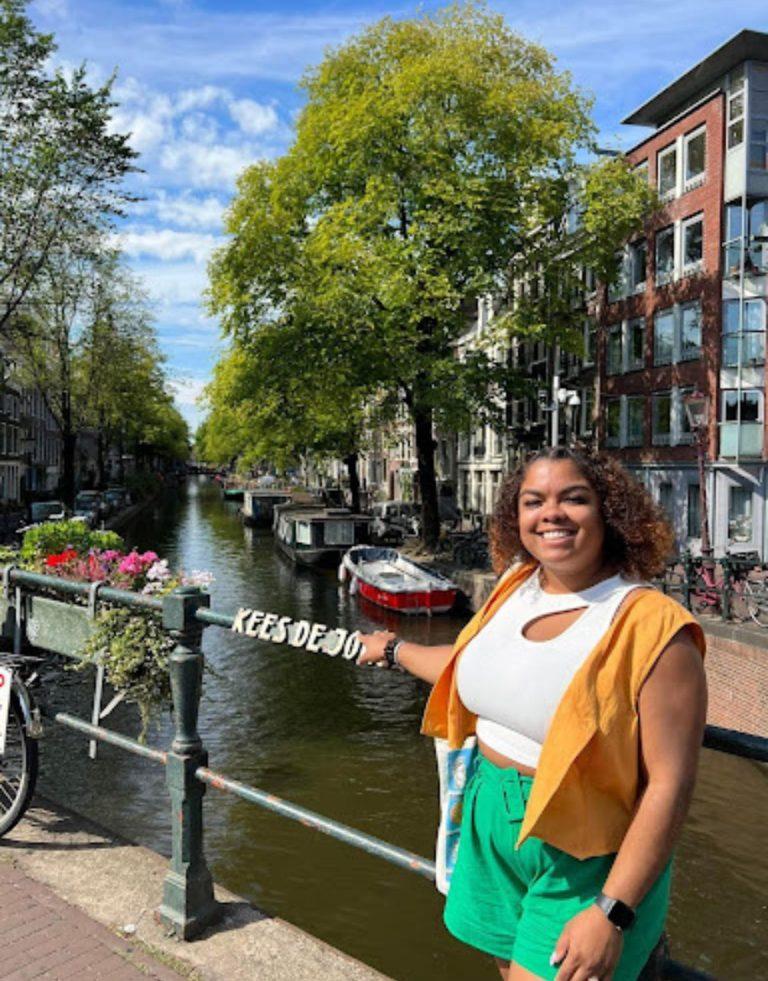 Una  Latina alegre posa a lo largo del pintoresco canal de Ámsterdam, rodeada de arquitectura tradicional holandesa y coloridas flores.