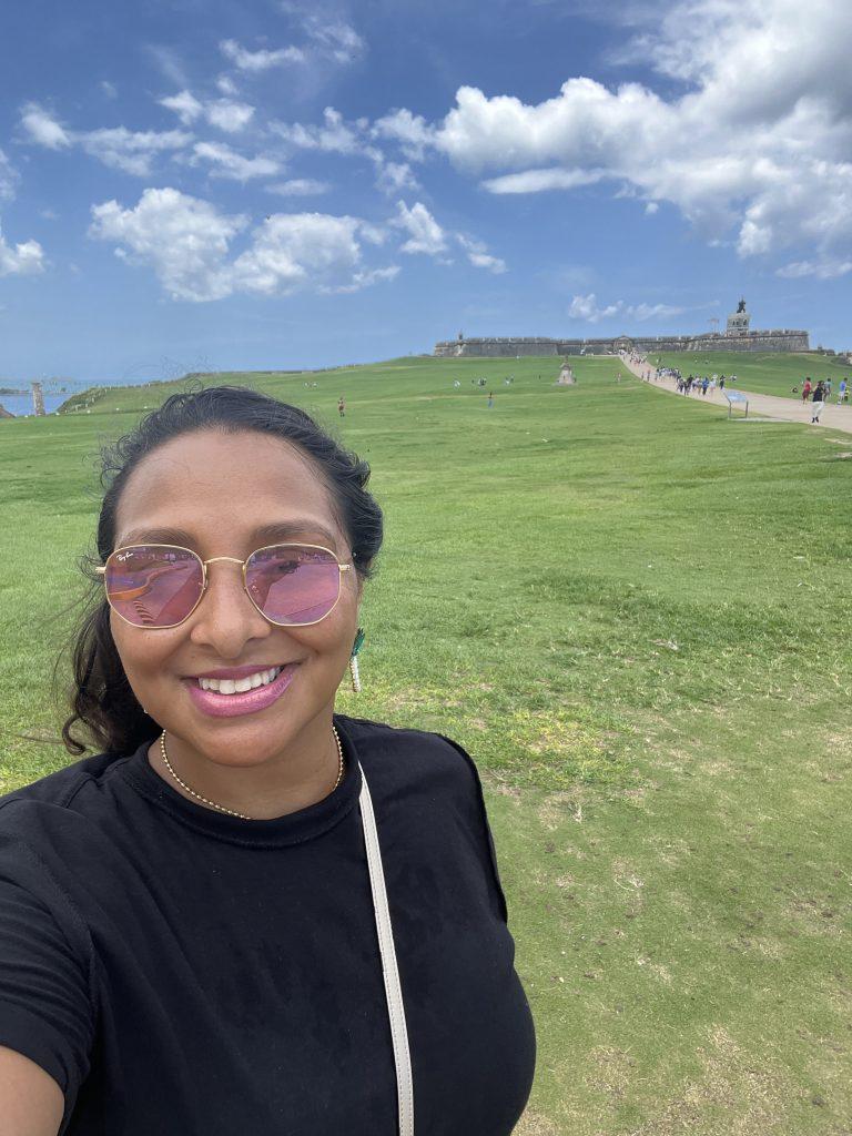 Latina traveler smiling in front of El Morro in San Juan, Puerto Rico during a 2022 visit. Make sure to stop by during Bad Bunny's Residency
