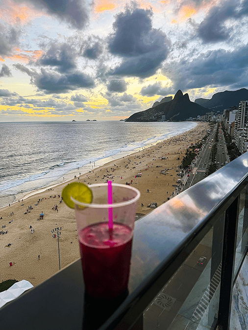 Sunset view of Ipanema Beach and Dois Irmãos mountains from the balcony of an oceanfront Airbnb in Ipanema, Rio de Janeiro, with a caipifruta drink in the foreground, March 2026.