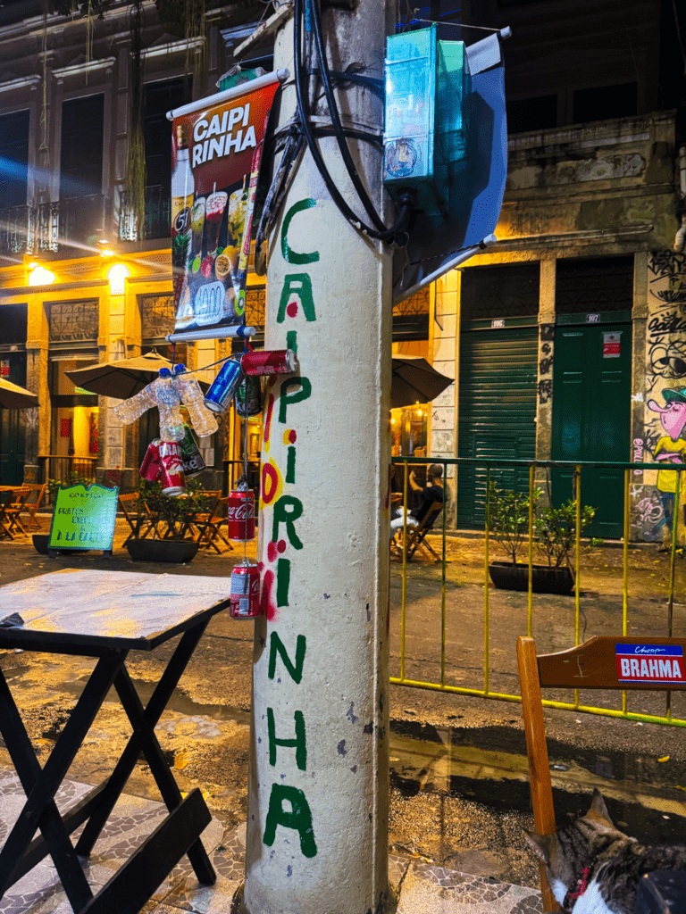 Where to stay in Rio: CAIPIRINHA painted vertically on a column in the Lapa neighborhood at night with bar seating, Brahma signage, and classic Lapa architecture in the background