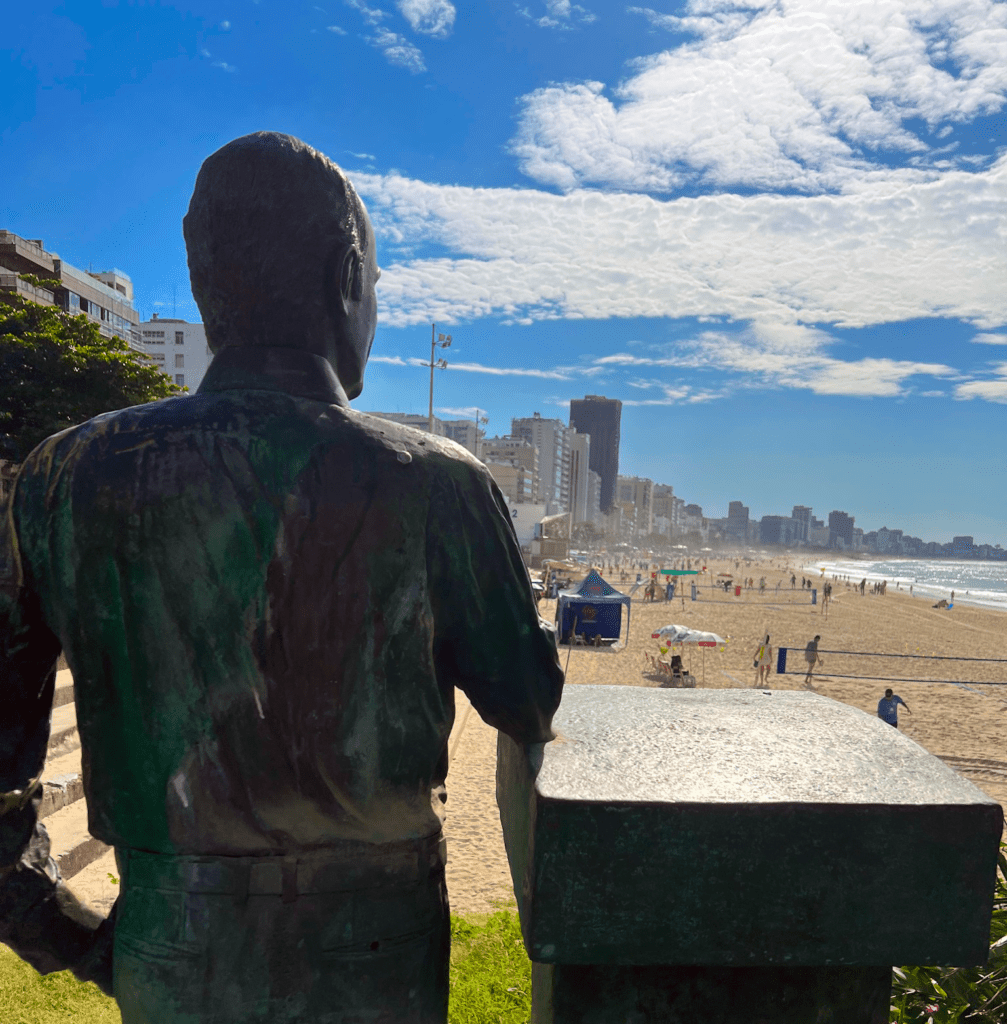 Where to stay in Rio: the Tom Jobim statue at the western end of Leblon beach in Rio de Janeiro looking east toward Ipanema beach with dramatic clouds and the coastline extending toward Pedra do Arpoador